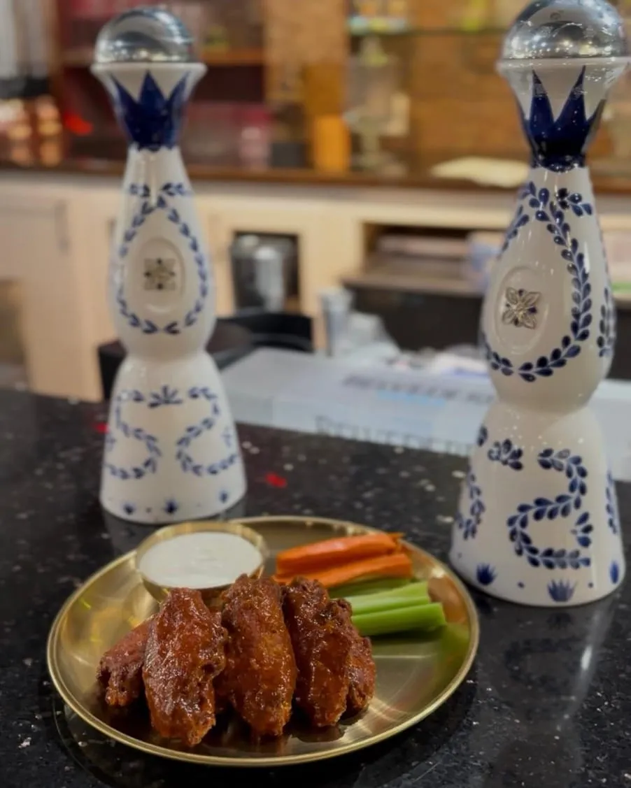 Glazed tikka wings with carrot and celery sticks and a dip, on the bar between two tequila bottles.